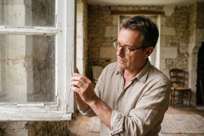 Homme inspectant le cadre abîmé d'une vieille fenêtre en bois dans une maison ancienne en pierre